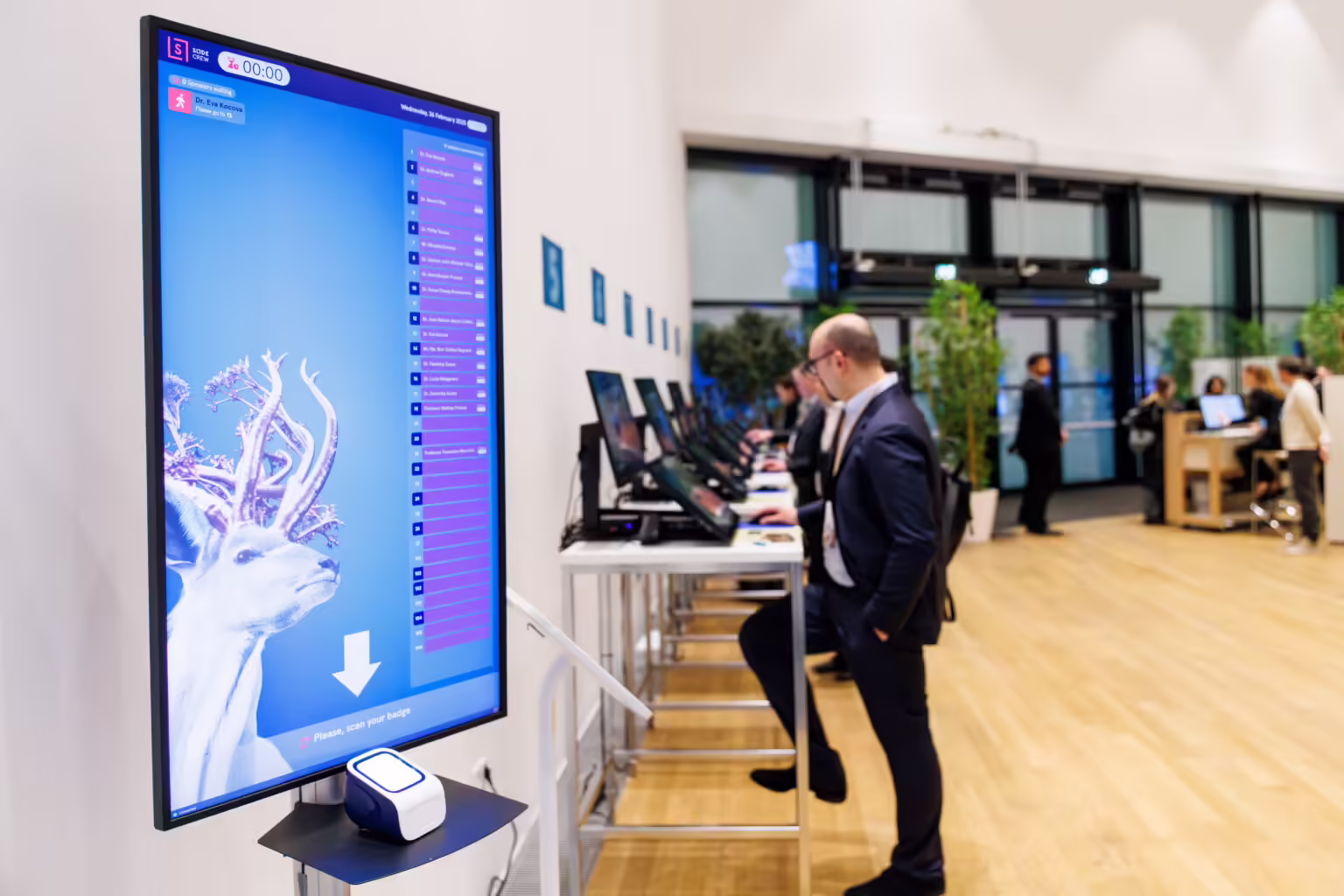 Slidecrew speaker queue display with built-in QR code scanner, presentation lineup; self-service speaker check-in stations visible in the background.